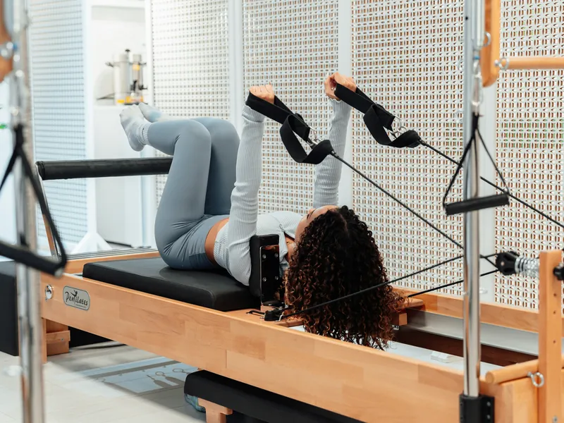 Ragazza sdraiata sul Reformer che tira le cinghie con le braccia, tra molle e telaio in legno